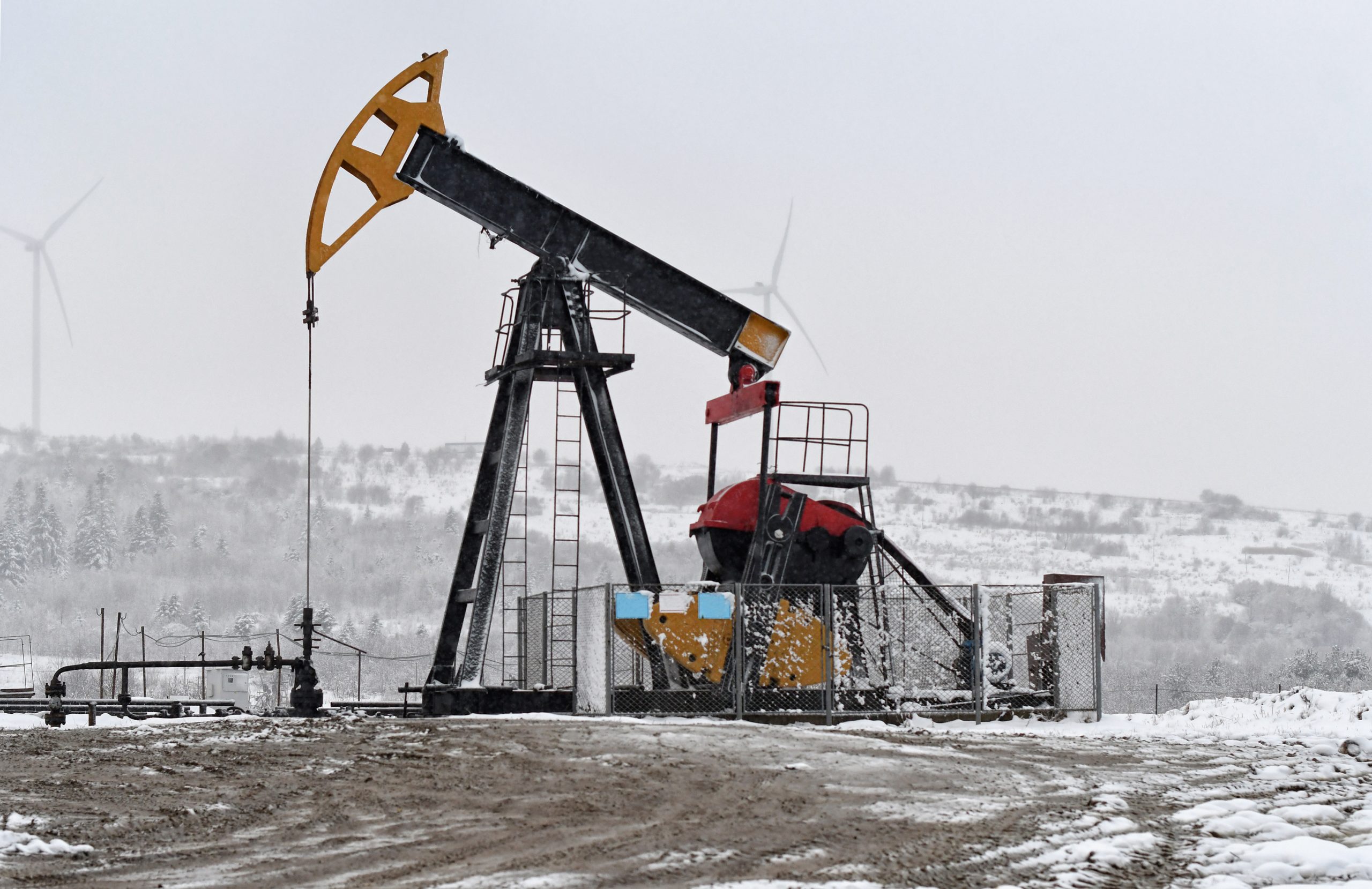 Oil pump. Oil and gas industry equipment. Oil field pump jack and oil refinery in the winter with snow, wind turbine mountains and forest in background